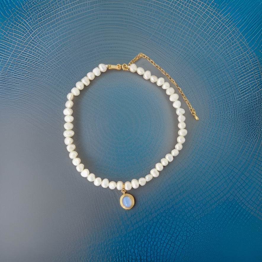 This is a white pearl necklace with a gold plated chain followed by a baby blue mini circular stone pendant plated in gold hanging at the bottom of the pearl necklace. The pearls go all around the necklace.